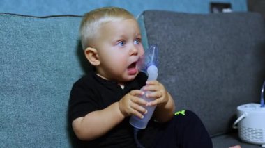Küçük bir çocuk kanepede oturur ve astım spreyiyle nefes alır. Çocuklar için terapi amaçlı solunum. Çocuklar için solunum maskesi.