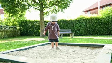 Yazın çocuk parkındaki kum havuzunda oynayan 2 yaşında bir çocuk. Yeni yürümeye başlayan çocuk sokakta kum kazıyor. Dina dışarıda oynuyor. Bir erkek için günlük kıyafetler. Tişörtlü ve şortlu çocuk.