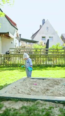 Panama şapkalı, kot tulumlu, kum havuzunda oynayan küçük bir çocuk. Güneşli yaz gününde küçük bir çocuk halk parkında oynuyor. Çocuk dışarıda yürüyor..