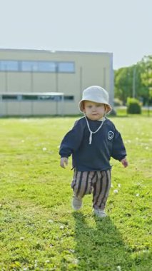 Kafasında panama şapkası olan 2 yaşındaki bir çocuk şehir parkında yeşil çimlerde koşuyor. Yazın yürüyüşe çıkmış küçük bir çocuk. Çocuklar için şık kıyafetler. Pantolonlu bir çocuk ve sokakta bir bluz..