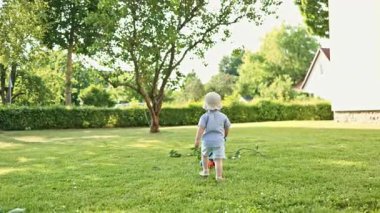 Beyaz saçlı, 2 yaşında şortlu ve tişörtlü bir çocuk bir yaz günü çimlerin üzerinde koşuyor. Parkta yürüyüş yapan şık kıyafetli bir çocuk. Bebek koşuyor. Kendi kendini geliştirme ditini.