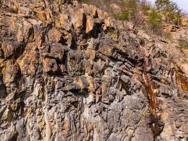 Tepenin yamacında çeşitli şekil ve renklerde kaya oluşumu görülüyor. Güneş ışığı taşların dokusunu vurgular. Ağaçlar açık bir günde kayaların etrafında büyür..