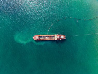 Büyük bir kargo gemisi derin mavi sularda demirli. Malzemeleri yükleme ve boşaltma için konumlandırıldı. Yakınlarda daha küçük teknelerin dolaştığı görülüyor. Güneş berrak bir gökyüzünde parlıyor.