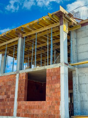 İşçiler inşaat alanında tuğladan duvarları ve iskelesi olan bir yapı inşa ediyorlar. Sahnede güneşli bir günde sütunlar, kirişler ve gökyüzü görülüyor..