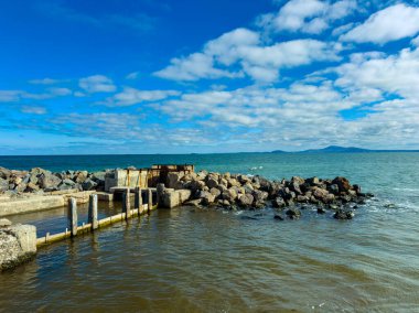 Deniz kıyısında bir yer iskelesi olan su ve kayaları gösteriyor. Dalgalar taşlara çarpıyor. Açık bir gökyüzü bulutları dağıttı. Bu sahne doğayı kıyıdan yakalar..