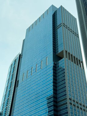 A tall glass skyscraper reaches upward against a cloudy sky in a vibrant city. Nearby buildings add to the skyline.