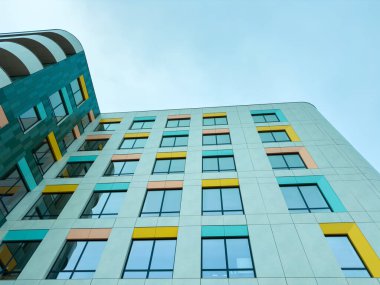 Looking up at a modern building that features colorful windows. The structure is made of various materials, set against a cloudy sky in an urban environment.
