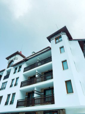 Birkaç balkonu olan yüksek bir bina sessiz bir alanda duruyor. Balkonlar boş ve gökyüzü bulutlu. Kış mevsiminde kar hafifçe yağar, manzaraya biraz doğa katar..