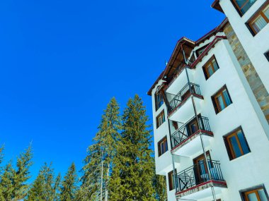 People enjoy the sun outside a white building with balconies. Tall green pine trees stand nearby. The sky is blue with clear weather on a sunny day in a mountain location.