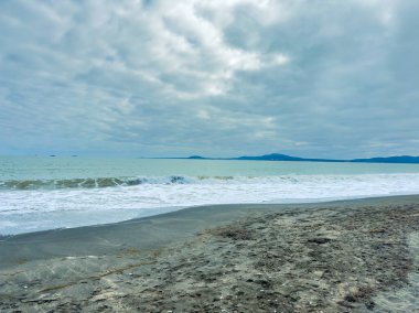 Dalgalar bulutlu bir gökyüzünün altında kumlu kumsalda yuvarlanıyor. Su ufukta görünen uzak tepelere kadar uzanıyor. Sahne, sahil boyunca doğanın bir anını yakalar..