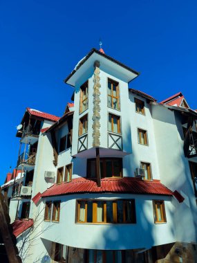 Yüksek bir bina açık mavi gökyüzünün altında birden fazla pencere ve kırmızı bir çatı gösteriyor. Gündüz vakti ağaçlar ve bölgedeki diğer evlerle çevrilidir..