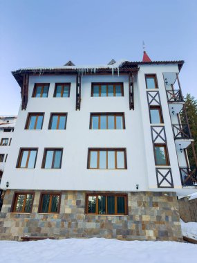 Beyaz duvarları ve karla çevrili taş zemini olan bir dağ kulübesi. Geniş pencereleri ve balkonları var. Buz saçakları açık mavi gökyüzünün altında asılı..