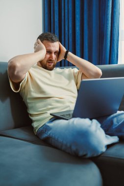 Bir adam elleri başının üstünde bir kanepede oturur ve dizüstü bilgisayara bakar. Rahat bir odada çalışırken stresli ya da bunalmış görünüyor..