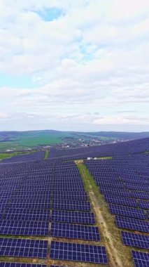 Güneş panelleri geniş bir alanı kırsal bir ortamda kaplıyor. Sahne, arka planda gündüz vakti açık bir gökyüzünün altındaki yeşil tepeleri ve tarım arazilerini gösteriyor..