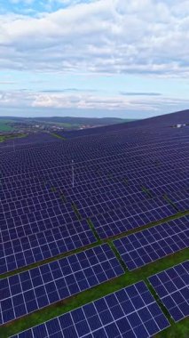 Güneş panelleriyle dolu büyük bir alan bulutlu mavi bir gökyüzünün altında görülür. Paneller kırsal alanda sıra sıra dizilmiştir..