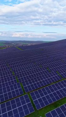 Bir güneş paneli çiftliği, birçok sıra güneş paneli olan yeşil bir araziye yayılır. Gökyüzündeki bulutlarla ortam aydınlık. Bakış açısı yenilenebilir enerji üretiminin ölçeğini yakalıyor.