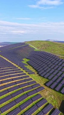 Güneş panellerinden oluşan bir alan, sıraların arasında yeşil çimlerin büyüdüğü bir yamaçta bulunuyor. Rüzgâr türbinleri arka planda bulutlu mavi bir gökyüzünün altında duruyor..