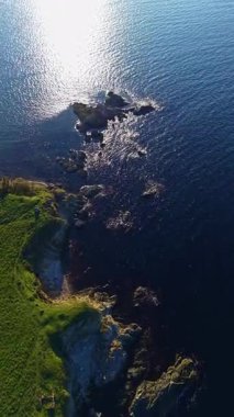 Su, yeşil toprakları ve uzak tepeleri olan kayalık bir sahilin yakınındaki güneş ışığını yansıtır. Sahne, açık bir günde, insanların olmadığı doğal güzellikleri gösteriyor..