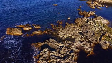 Yumuşak dalgaların sahili okşadığı derin mavi denizden kayalık kayalıklar yükselir. Akşam ışıklarının ılık renkleri suyun üzerinde dans ederek huzurlu bir atmosfer yaratıyor..