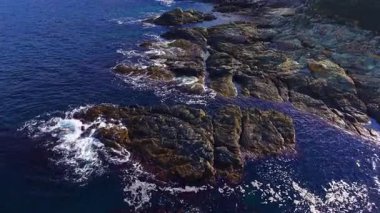 Derin mavi sulardan sert taşlar yükselir. Yumuşak dalgalar yüzeylerine vurur. Parlak bir güneş sahili aydınlatıyor, barışçıl ve doğal bir manzara yaratıyor..