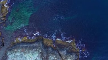 Engebeli kayaların sakin manzarası derin mavi okyanusla buluşur. Yumuşak dalgalar kıyıya vurur. Deniz kenarında sakin bir öğleden sonra güneş ışığı yüzeyde dans eder..