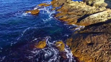 Dalgalar engebeli kıyı şeridine hafifçe vurarak kayaların üzerindeki ışık ve gölgenin etkileşimini ortaya çıkarır. Huzurlu an, sahil boyunca doğanın güzelliğini yakalar..