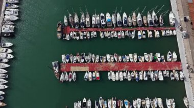 Birçok tekne marina rıhtımında dizilmiştir. Su temiz ve yeşil görünüyor. Güneş ışığı, farklı boylarda ve şekillerde tekneler göstererek sahneyi aydınlatıyor..