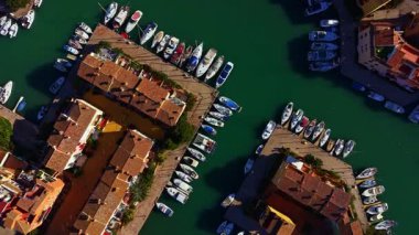 Several boats are arranged neatly at the docks next to buildings. The scene shows a busy harbor in a coastal town during the day under clear skies.
