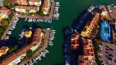 A marina shows multiple boats moored alongside residential buildings. The scene includes blue water and a few parked cars. Sunlight shines down on the area, highlighting the boats.