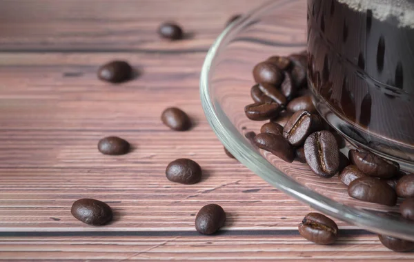 Close up transparent glass hot espresso cup filled with fresh organic coffee beans and roasted beans on wooden floor and copy space