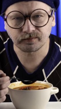 Tom yer elması yiyen bıyıklı bir adamın yakın çekimi. Karidesli kaşık. Kalın kırmızı çorba. Dikey video. Dikey olarak. Mavi arkaplan.