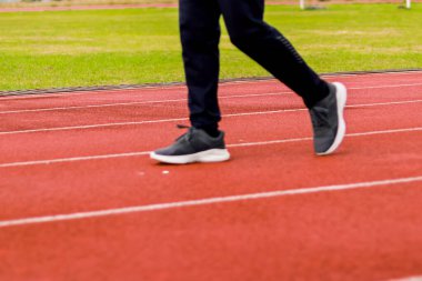 Atletik pantolon ve spor ayakkabılarla beyaz şeritli kırmızı bir pistte yürüyen birinin bacaklarının yakın çekimi..