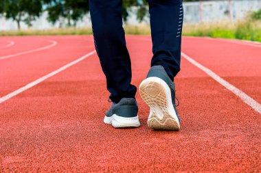 Kırmızı bir koşu pistinde yürüyen, spor ayakkabıları ve koyu renk pantolonlar giyen bir insanın bacak ve ayaklarının yakın görüntüsü..