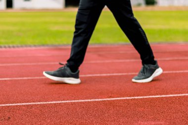 Kırmızı bir atletizm pistinde yürüyen, siyah spor ayakkabı ve siyah pantolon giyen birinin bacakları ve ayaklarının yakın çekimi..