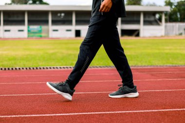 Kırmızı bir atletizm pistinde arka planda bir stadyumla yürüyen bir insanın bacak ve ayaklarının yakın çekimi..