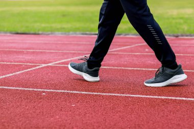 Kırmızı bir atletizm pistinde beyaz şerit işaretleriyle yürüyen bir insanın bacakları ve ayaklarının yakın çekimi..
