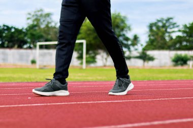 Kırmızı bir pistte yürürken, arka planda yeşil bir tarla ve ağaçlarla bir insanın bacaklarının yakın plan görüntüsü..