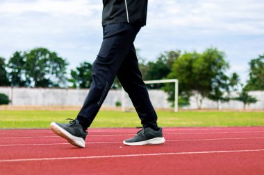 Kırmızı bir atletizm pistinde yürürken çimenli bir tarlası ve ağaçları olan bir insanın bacaklarının yakın çekimi..