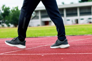 Kırmızı bir atletizm pistinde yürüyen, koyu renk spor pantolonları ve spor ayakkabıları giyen bir insanın bacak ve ayaklarının yakın çekim görüntüsü..
