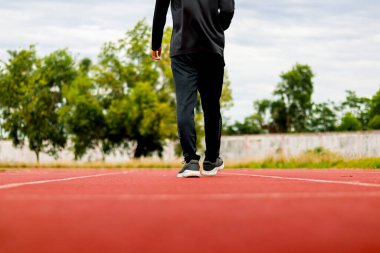 Yeşil ve bulutlu gökyüzü ile çevrili kırmızı bir pistte yürüyen atletik giysili bir insan..