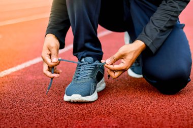 Kırmızı bir atletizm pistinde koşan bir ayakkabının bağcıklarını bağlamak için çömelmiş birinin yakın çekimi..