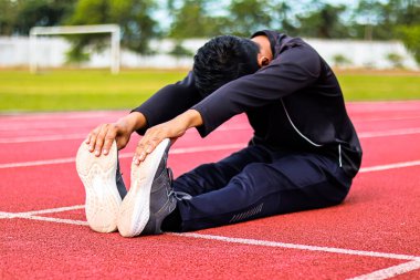 Bir adam koşu pistinde oturur, ayak parmaklarına uzanarak dizlerini uzatır..