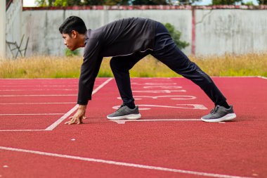Atletik giyimde kararlı genç bir adam kırmızı koşu pistinin başlangıç çizgisinde koşmaya hazır..