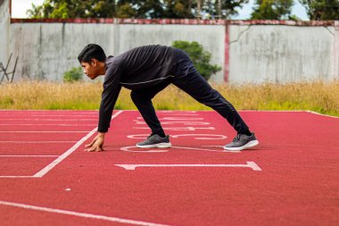 Kararlı genç bir atlet kırmızı bir atletizm pistinin başlangıç çizgisinde çömeliyor..