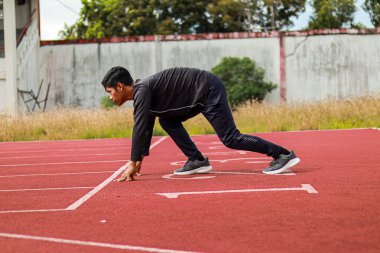 Siyah eşofmanlı genç bir erkek atlet kırmızı bir koşu pistinin başlangıç çizgisinde yarışa hazırlanıyor..