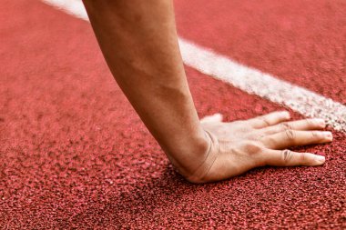Kırmızı bir atletizm pistinin başlangıç çizgisinde hareketlenmeye hazır bir koşucunun elinin yakın çekimi.