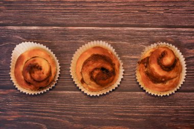 Üç taze fırınlanmış tarçınlı tatlı çörek. Kağıt kaplamalarda sunuluyor. Koyu, ahşap bir masa arkasında..