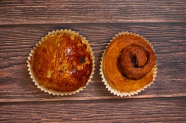 İki yeni pişmiş hamur işi, bir kuru üzümlü çörek ve bir tarçınlı rulo, ahşap bir yüzeyde sunuluyor..