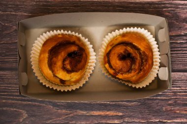 Kağıt bardaklarda pişirilmiş iki tatlı tarçın rulosu, sade ahşap bir yüzeyde basit bir karton kapta sunuluyor..