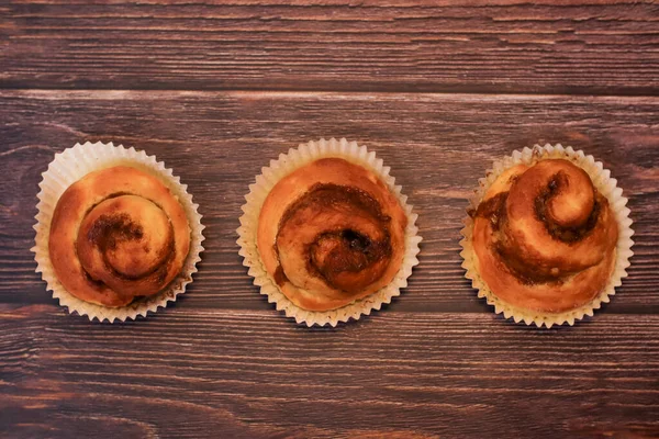 Üç taze fırınlanmış tarçınlı tatlı çörek. Kağıt kaplamalarda sunuluyor. Koyu, ahşap bir masa arkasında..
